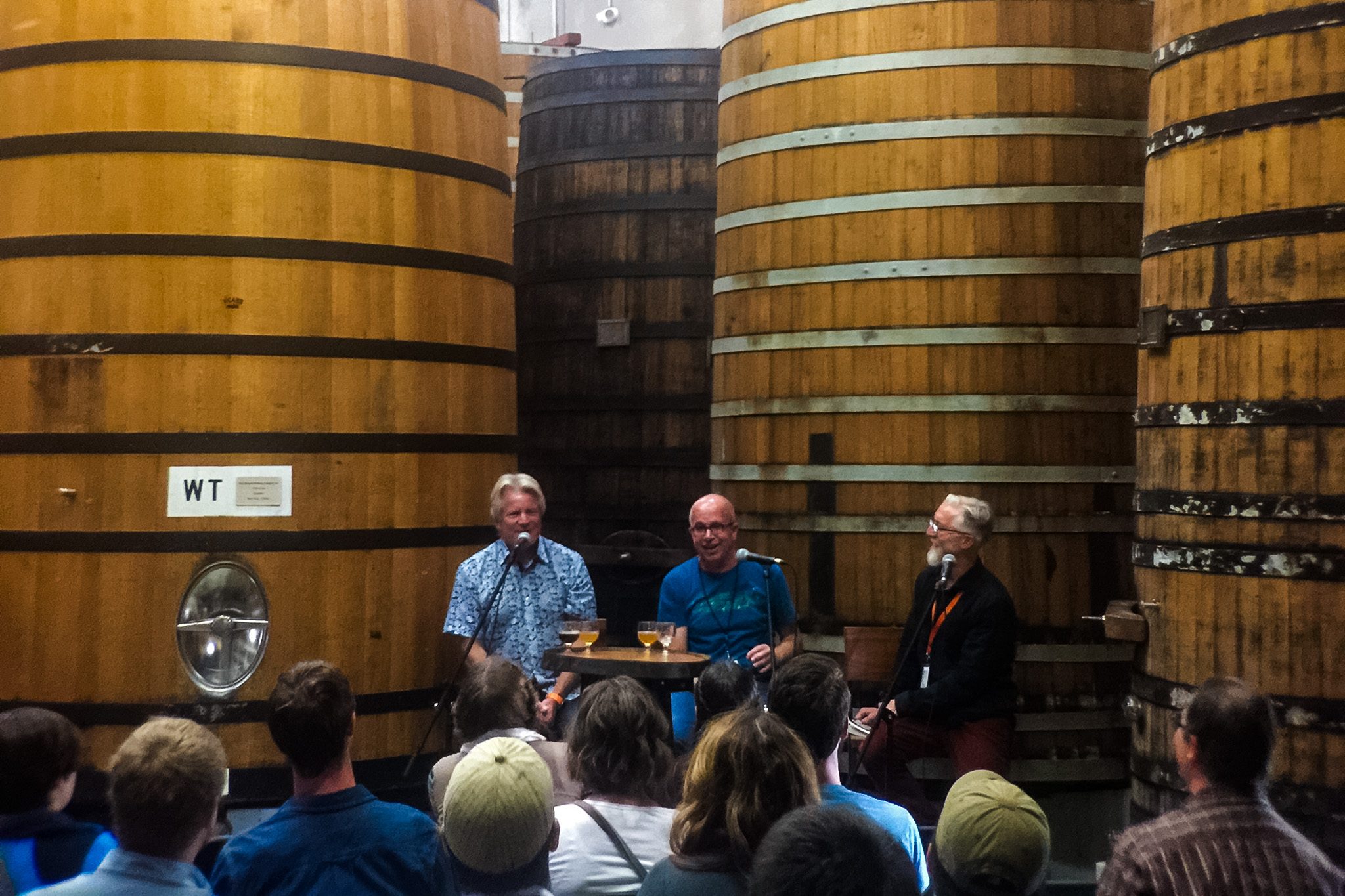 three men with microphones speaking in front of giant fermentation barrels to a room of people